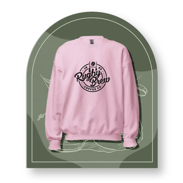 A light pink crewneck rugby sweatshirt with a black circular 'Rugby Brew Coffee Co' logo printed on the chest, presented on a dark olive green arched graphic background.